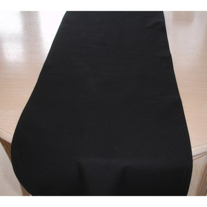 May include: A black table runner made of fabric. The runner is lying flat on a light wood table.