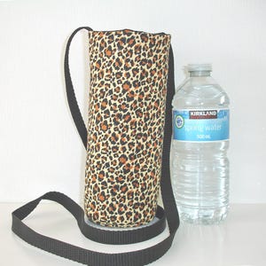 May include: A cylindrical fabric bottle carrier featuring a tan, black, and brown leopard print pattern with a long black adjustable strap. The carrier is positioned next to a clear plastic Kirkland Signature spring water bottle labeled 500 ml.