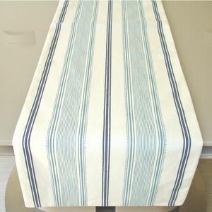 May include: A rectangular table runner featuring a cream-colored base with a repeating pattern of vertical stripes in varying shades of blue. The design includes thick dark navy lines paired with thinner, lighter teal pinstripes and dotted lines.