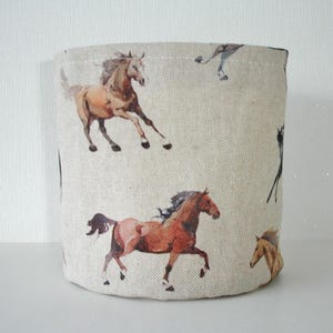 May include: A cylindrical fabric storage basket featuring a repeating pattern of watercolor-style horses in shades of brown, tan, and black against a light beige, textured linen-look background. The horses are depicted in various running poses.