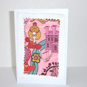 May include: A white greeting card featuring a rectangular fabric patch with a pink, yellow, and teal illustration. The design depicts a figure with yellow hair, a red dress, and a pink building, all outlined in black with visible stitching.