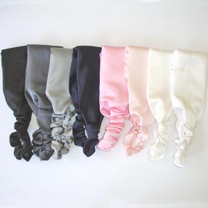May include: A collection of eight satin headbands arranged in a row, featuring a smooth finish and an elasticated gathered section at the base. The colors include black, charcoal gray, light gray, navy blue, soft pink, blush, cream, and white.