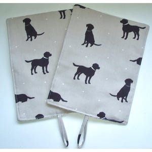 May include: Two beige oven mitts with black silhouettes of Labrador Retrievers printed on them. The mitts have a white polka dot pattern and a hanging loop.