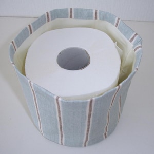 May include: A cylindrical fabric storage basket featuring a light blue and white striped pattern with thin brown vertical lines. The interior is lined with a solid off-white fabric, and the basket contains a single roll of white toilet paper.