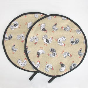May include: Two circular pot holders with a beige background, black trim, and handles. They feature a repeating pattern of black and white rooster illustrations with red details. These pot holders are designed for kitchen use.