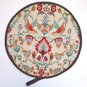 May include: Round, cream-coloured fabric trivet with a brown leather trim and a brown leather handle. The fabric features a repeating pattern of red, blue, and green stylised flowers and birds.