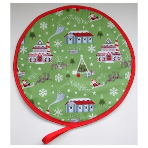 May include: Circular Christmas tree skirt with a green backdrop, showcasing a festive design of Santa's sleigh, reindeer, houses, snowflakes, and Christmas trees. It has a red border and a red handle.