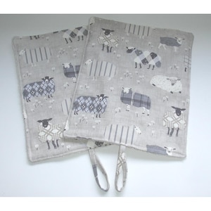 May include: Two grey oven mitts featuring a sheep design. The sheep are shown in different poses and wearing plaid and striped patterns. Each mitt has a loop for hanging.