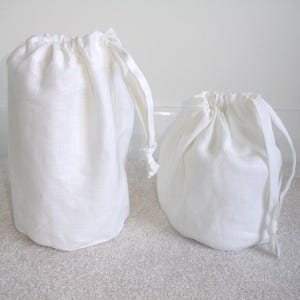 May include: Two white linen drawstring bags. One is tall and cylindrical, the other is shorter and rounded. Both bags have a gathered top with a drawstring closure. The bags are made of a textured, natural-looking fabric.