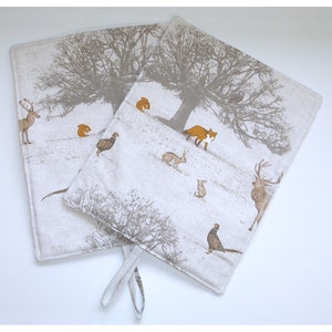 May include: Two beige linen pot holders with a woodland animal print. The print features a deer, fox, squirrels, rabbits, and pheasants. The pot holders have a loop handle.