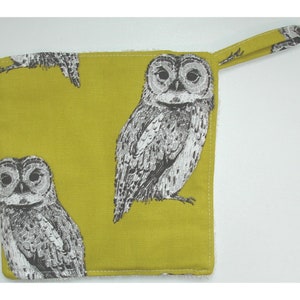 May include: A square, olive-green fabric item with a repeating pattern of black and white owl illustrations. It has a loop on one corner and a white, fluffy border. The item is likely a kitchen accessory.