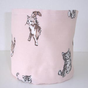 May include: A cylindrical fabric storage bin in a soft pastel pink color featuring a repeating pattern of various kittens in shades of brown, gray, and white. The kittens are depicted in different poses, including sitting and walking, across the exterior.