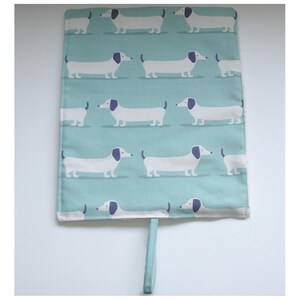 May include: A light blue fabric placemat with a white dachshund pattern. The placemat has a white fabric border and a white fabric loop handle.