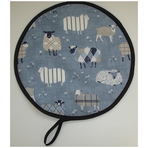 May include: A round blue pot holder with a black trim and a black handle. The pot holder has a pattern of white, black and grey sheep on a blue background.