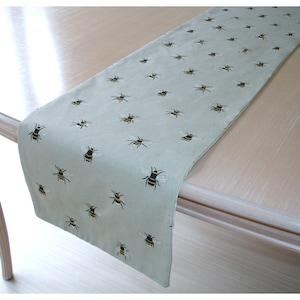 May include: A light green table runner with an embroidered bee pattern. The bees are black and yellow with white wings, scattered across the fabric. The runner is draped over a light-colored wooden table.