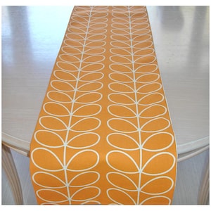 May include: An orange table runner featuring a white leaf design. The runner is rectangular and rests on a light-coloured table. The leaf pattern is symmetrical, with each leaf shape outlined in white.