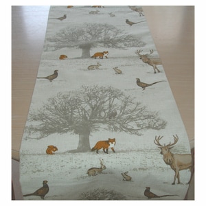May include: A beige table runner with a woodland pattern. The pattern features a large tree, deer, foxes, squirrels, and birds. The background is a light beige colour.
