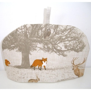 May include: A beige tea cozy with a woodland scene featuring a fox, a deer, a squirrel, an owl, and a rabbit. The cozy is made of linen and has a loop for hanging.