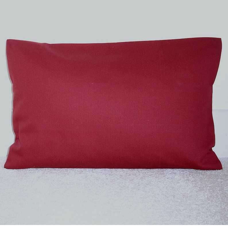 May include: A solid red rectangular cushion with a smooth, even surface. The cushion is lying flat on a white, fluffy surface.
