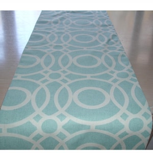May include: A rectangular table runner with a repeating white geometric pattern on a light blue background. The design features interlocking circles and lines, creating a modern look. The runner is made of fabric and is suitable for home decor.