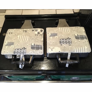 May include: Two grey and white patterned waffle iron covers with sheep designs. The covers are on a black stovetop.