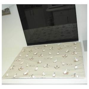 May include: A rectangular kitchen mat in beige, patterned with numerous ducks in white, brown, and grey. The mat is placed on a white surface, with a dark appliance reflected in the background. The mat's design is repeated across its surface.