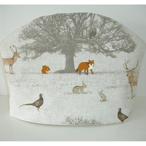May include: A tea cozy with a woodland scene. The cozy features a large tree with an owl perched in its branches. Various animals, including deer, a fox, rabbits, a squirrel, and pheasants, are scattered across the snowy landscape.
