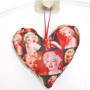 May include: A red heart-shaped cushion featuring a collage of images of Marilyn Monroe. The cushion has a red ribbon attached for hanging.