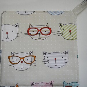 May include: A beige fabric pot holder with a repeating pattern of cartoon cats wearing glasses. The cats have different colored glasses and facial expressions.