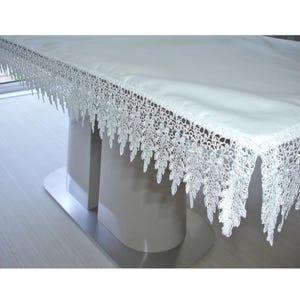 May include: A white rectangular tablecloth featuring a wide, intricate lace border with a detailed floral and leaf pattern. The fabric is smooth and opaque, draping over the edges of a table, with the lace trim hanging down in a decorative fringe.