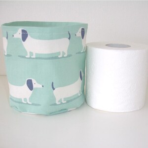 May include: A cylindrical fabric storage basket featuring a repeating pattern of white dachshunds with blue ears on a light mint green background, positioned next to a standard roll of white toilet paper for a clear visual size comparison.