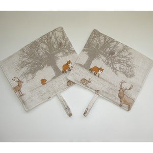 May include: Two beige oven mitts with a woodland scene design. The mitts feature a brown tree with a brown owl, a red fox, a deer, a squirrel, and a rabbit. The mitts have a brown loop handle.