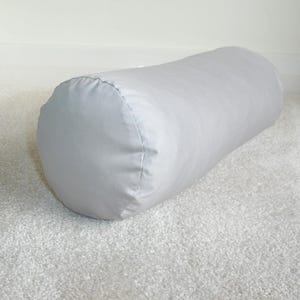 May include: A light gray cylindrical bolster pillow. The pillow is made of a smooth fabric and is approximately 20 inches long. The ends are rounded and sewn with a visible seam. The pillow is resting on a white carpet.