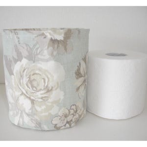 May include: A cylindrical fabric storage basket featuring a light blue and beige floral pattern, positioned next to a standard white roll of toilet paper. The basket has a soft, unstructured design with a visible woven texture throughout the fabric.