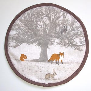 Peut inclure: Un tapis en tissu rond avec une bordure cousue marron présentant une scène imprimée d'un grand arbre gris, d'un hibou perché, d'un petit écureuil orange, d'un renard orange debout et d'un lapin brun sur un fond gris clair texturé.