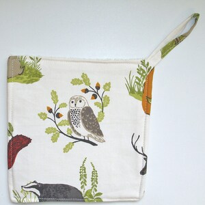 May include: White cotton pot holder with a woodland theme. The design features an owl, a badger, a deer, and acorns. The pot holder has a loop for hanging.