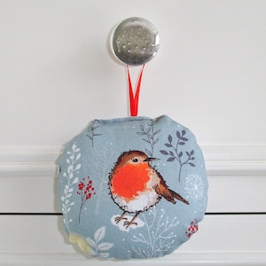 May include: A round, light blue fabric ornament featuring a detailed illustration of a robin with a bright orange breast. The ornament is decorated with white and grey floral patterns and red berries. A red ribbon is attached to a clear, round knob.