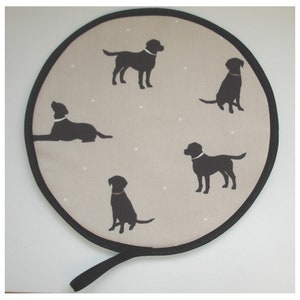 May include: A round, black and beige fabric trivet with a black handle. The trivet is decorated with five black silhouettes of Labrador Retrievers.
