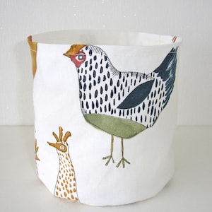 May include: A white fabric storage bin with a whimsical chicken design. The bin features a large chicken illustration with black spots, a red comb, and green accents. A smaller chicken illustration is also visible.