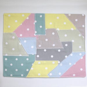 May include: A rectangular patchwork placemat with a variety of pastel-colored fabric pieces. The pieces are in shades of yellow, pink, blue, gray, and green, each with white polka dots. The edges are stitched with white thread.