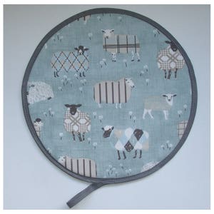 May include: A round, blue and white fabric pot holder with a black trim and a black hanging loop. The pot holder features a repeating pattern of sheep with different coloured wool.