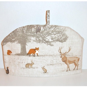 May include: A beige tea cozy with a woodland scene featuring a fox, deer, owl, squirrel, and rabbits. The cozy has a loop handle for easy use.