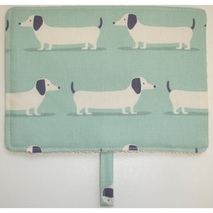 May include: A teal blue kitchen towel with a white border and a repeating pattern of white dachshunds with black ears and noses. The towel has a loop handle.