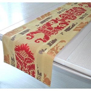 May include: A gold table runner with a red and brown floral pattern. The runner has a damask design with a repeating pattern of stylised flowers and leaves.