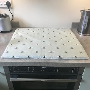 May include: A pale blue rectangular hob cover adorned with a repeating pattern of black, white, and yellow bees. The cover rests on a black oven. A stainless steel kettle and blender are visible in the background.