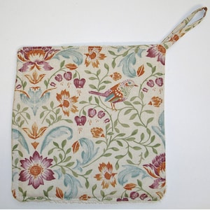 May include: A cream-coloured pot holder with a floral pattern in shades of orange, red, green, and blue. A small bird with brown, orange, and green feathers is perched on a branch in the centre of the pot holder. The pot holder has a loop handle.