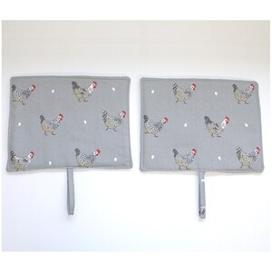 May include: Two grey pot holders featuring a repeating pattern of chickens and white egg shapes. Each has a hanging loop. The chickens are grey, black, and white with red combs.