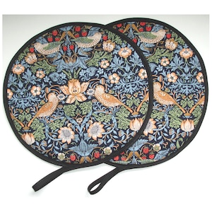 May include: Two circular pot holders with a black trim and a black loop handle. The fabric displays a dark blue background with a floral and bird design in green, orange, and light blue. The pattern includes birds, flowers, and strawberries.