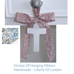 May include: A white cross suspended by a floral ribbon bow. The ribbon displays a pink and red floral design. The text at the bottom reads "Choice Of Hanging Ribbon Handmade - Liberty Of London."