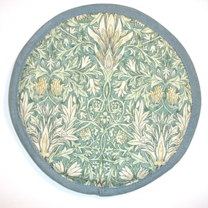 May include: Round, floral-patterned trivet with a light blue border. The trivet features a detailed design of green and cream-colored leaves and flowers. The overall aesthetic is reminiscent of vintage botanical illustrations.